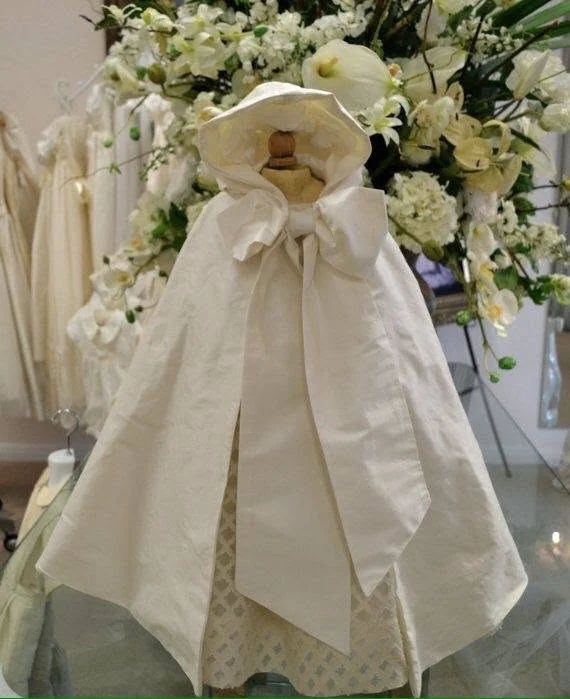 Capa de bautizo marfil en maniquí, con lazo frontal y flores.